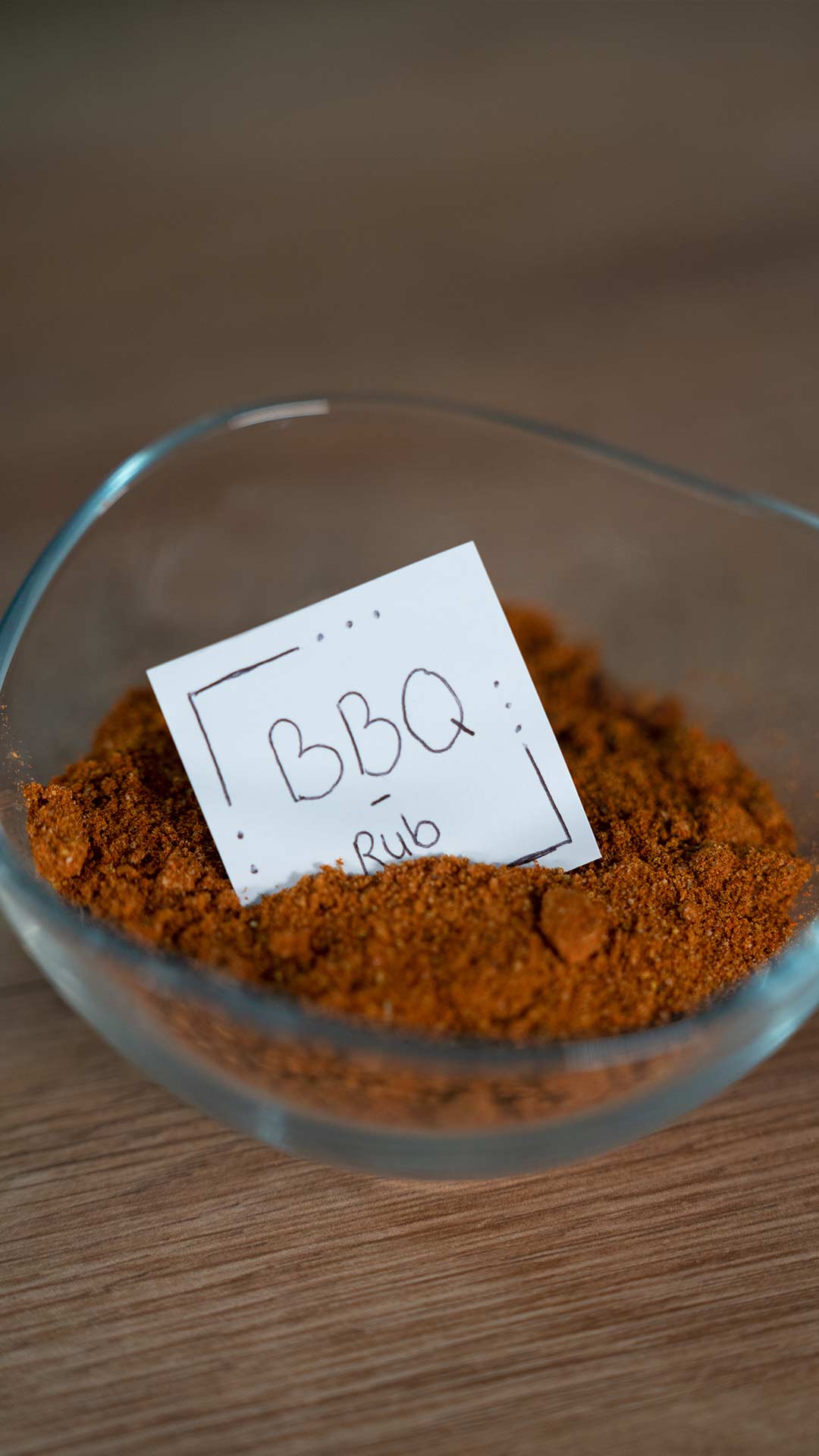 Auf einer Holzoberfläche steht eine Glasschale mit einer braunen BBQ-Gewürzmischung. In der Schale befindet sich ein kleines handgeschriebenes Etikett mit der Aufschrift „BBQ Rub“.
