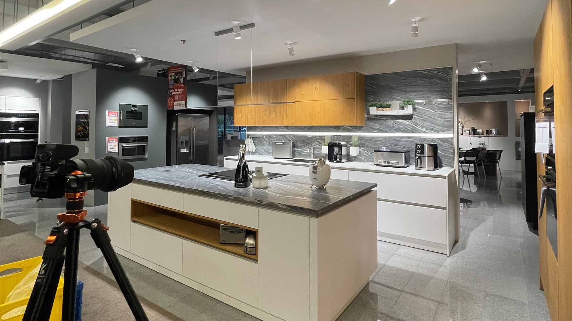 Moderner Küchenausstellungsraum mit Marmorinsel, Holzschränken, verschiedenen Geräten und einem Stativ mit einer Kamera im Vordergrund. Der Raum ist hell erleuchtet und in schlichtem, minimalistischem Design gehalten.