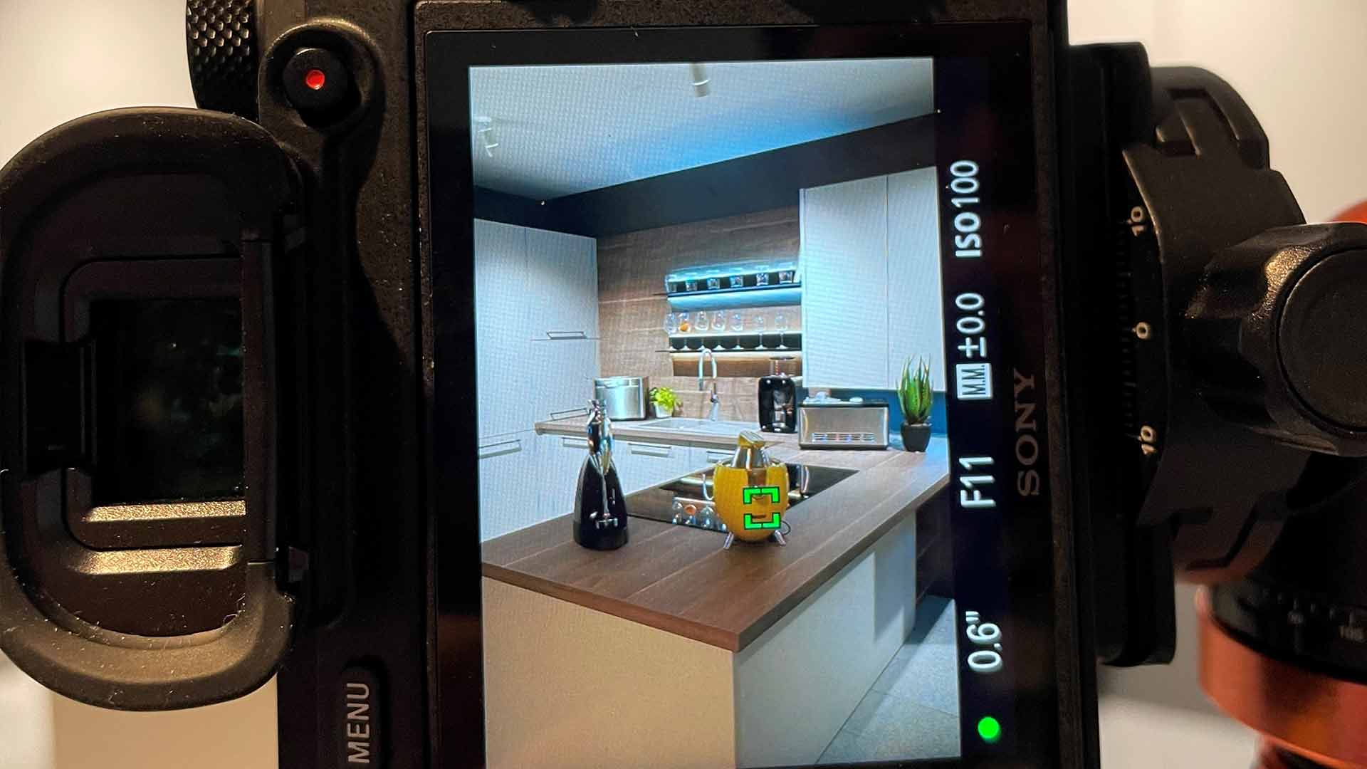 Auf dem Kamerabildschirm ist eine moderne Küche mit hellen Schränken, einer Holzrückwand und einer Theke mit Flaschen, einem Wasserkocher und einer Kaffeemaschine zu sehen. Die Kameraeinstellungen sind rund um das Bild sichtbar.