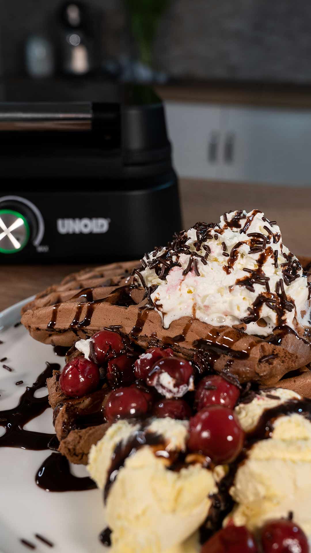 Ein Teller mit Schokoladenwaffeln, garniert mit Schlagsahne, Schokoladensirup, Kirschen und Kugeln Vanilleeis, im Hintergrund ein Waffeleisen.
