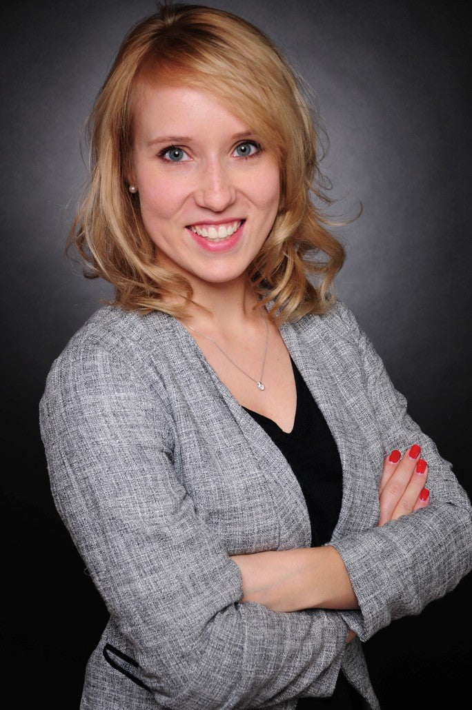 Eine Frau mit blonden Haaren, einem grauen Blazer und einem schwarzen Oberteil trägt, lächelt mit verschränkten Armen vor einem dunklen Hintergrund.