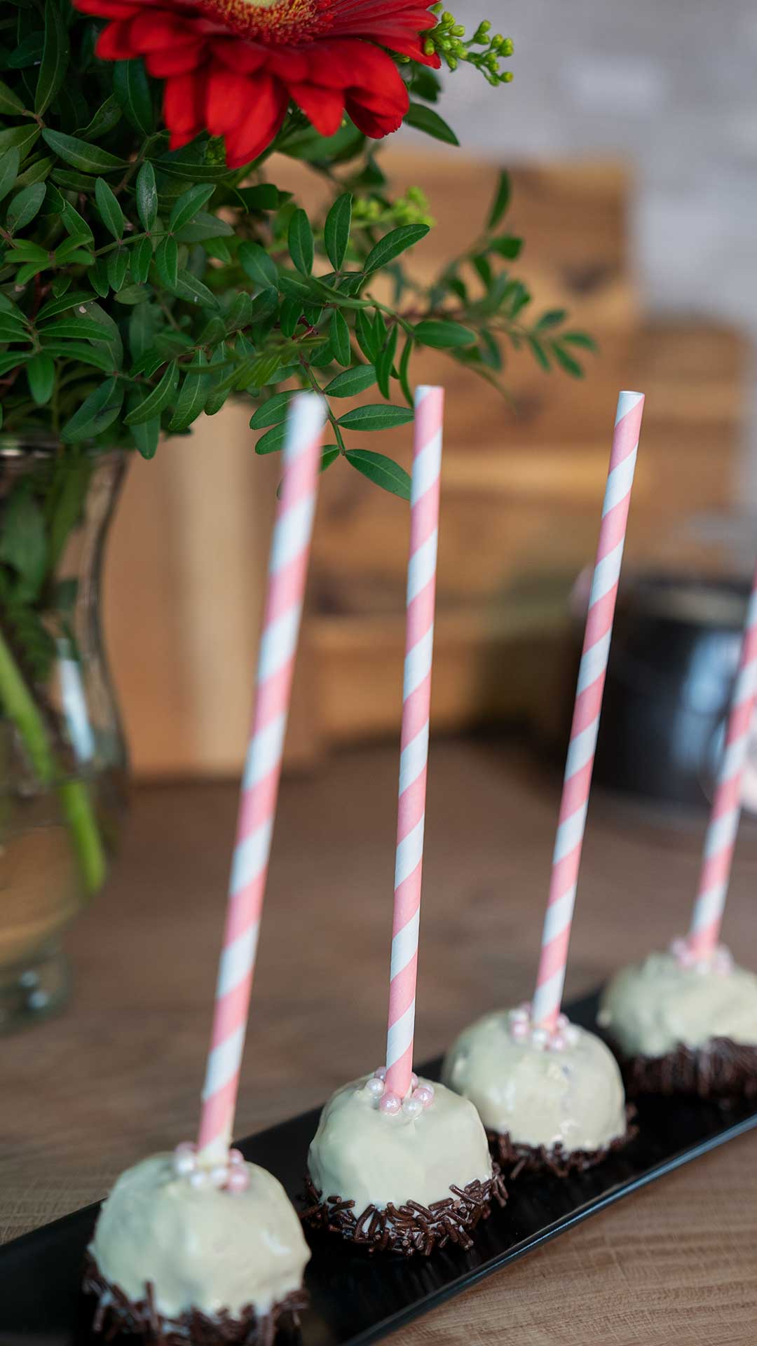 Vier Cake Pops mit weißem Zuckerguss, Schokostreuseln am Boden und rosa gestreiften Strohhalmen stehen aufrecht auf einem schwarzen Tablett. Im Hintergrund ist ein Strauß aus roten Blumen und grünen Blättern zu sehen.
