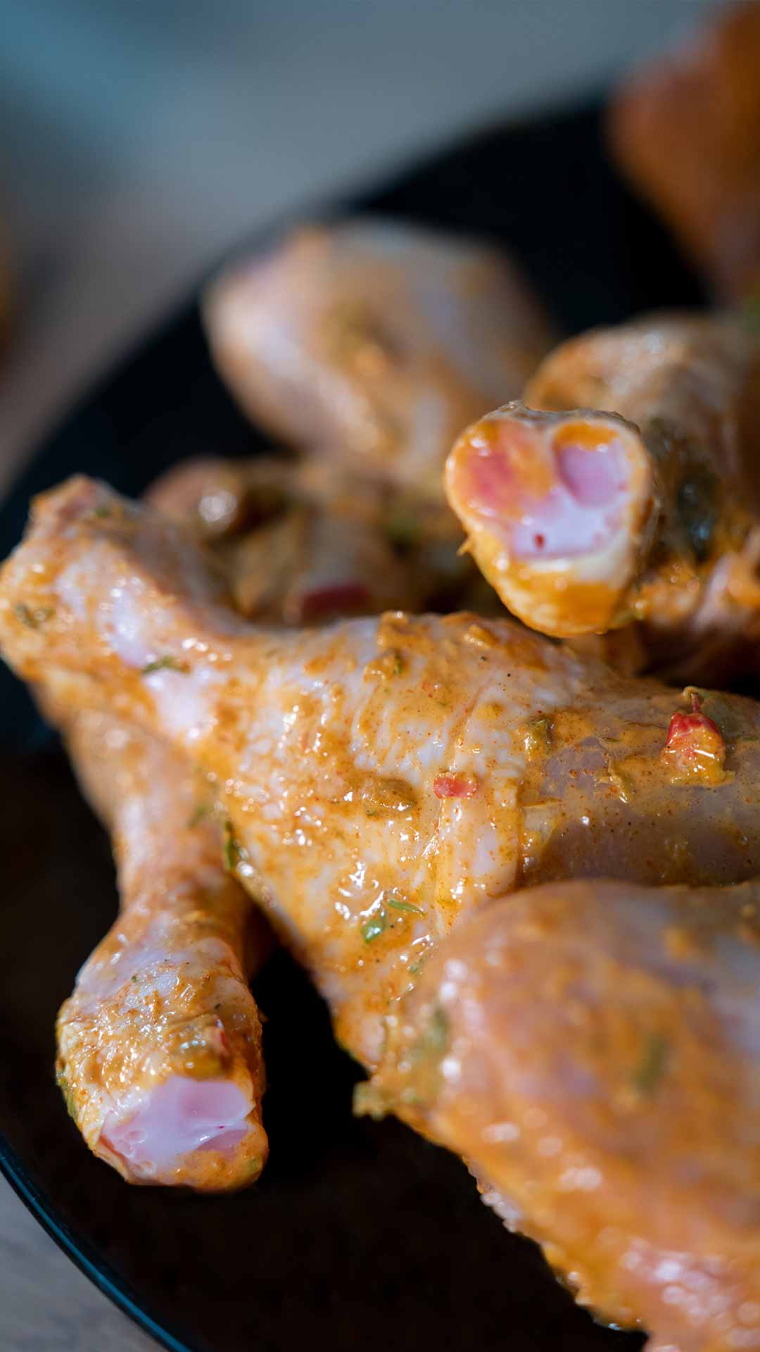 Nahaufnahme von rohen Hähnchenkeulen, überzogen mit Gewürzen und Kräutern, angerichtet auf einem dunklen Teller, mit sichtbaren Gewürzen und Knochenenden.