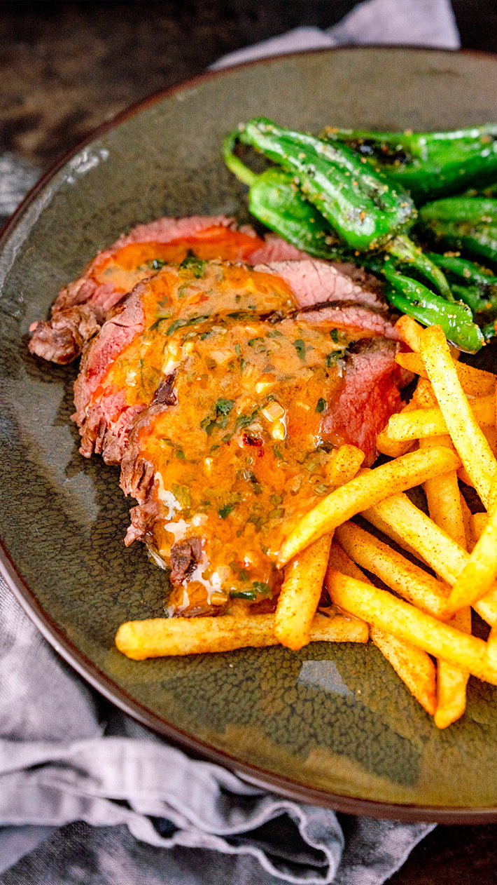 Geschnittenes Steak mit einer cremigen Kräutersauce, serviert mit goldbraunen Pommes Frites und sautierten grünen Paprikaschoten auf einem dunklen Teller.