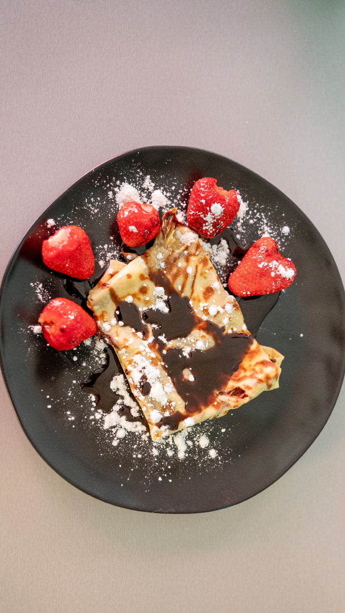 Ein schwarzer Teller mit einem gefalteten Crêpe, beträufelt mit Schokoladensoße, umgeben von ganzen und herzförmigen Erdbeeren und bestäubt mit Puderzucker.