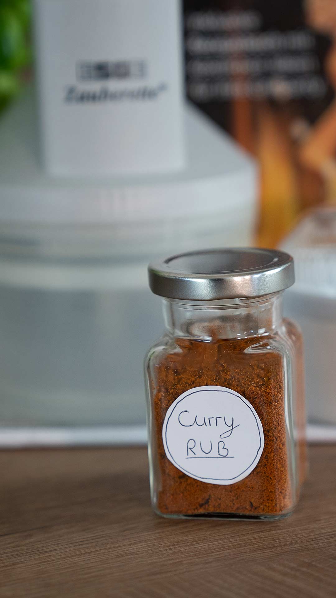 Ein kleines Glasgefäß mit silbernem Deckel, gefüllt mit rotbrauner Curry-Gewürzmischung, steht auf einer Holzfläche. Das Glas trägt ein handgeschriebenes Etikett mit der Aufschrift „Curry RUB“. Der Hintergrund ist unscharf.