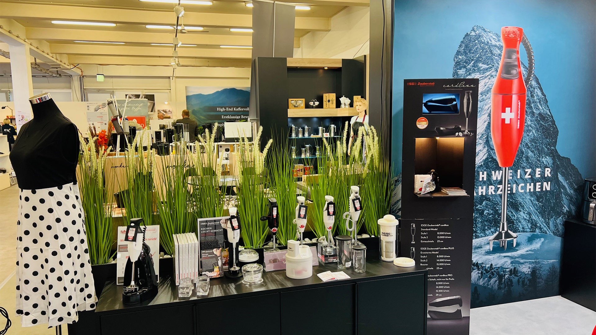 Eine moderne Ladenauslage mit Küchengeräten, einer Schaufensterpuppe in einem schwarz-weiß gepunkteten Kleid, grasähnlichem Dekor und einem großen Poster eines Schweizer Stabmixers vor einer Bergkulisse.