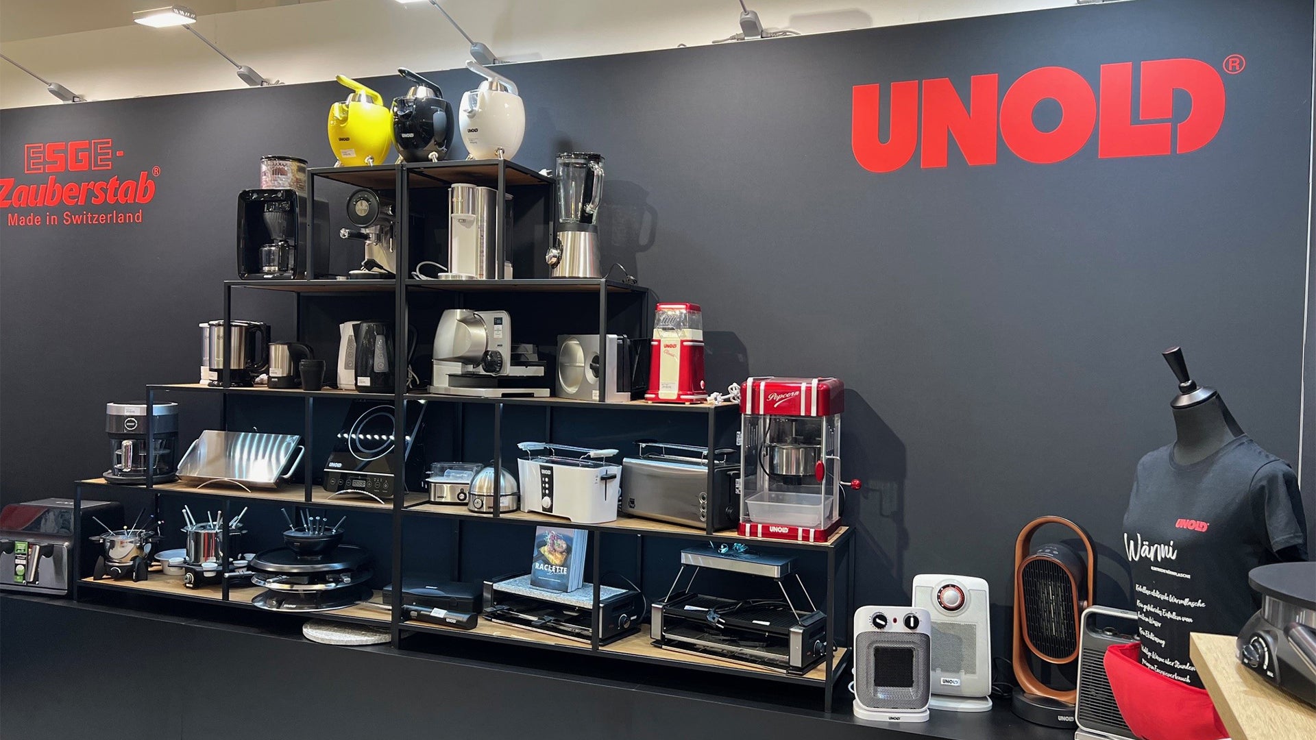 Ein Ausstellungsregal auf einer Messe mit verschiedenen Küchengeräten wie Mixern, Toastern, Kaffeemaschinen und Rührgeräten. Auf dunklem Hintergrund sind die Marken ESGE Zauberstab und UNOLD zu sehen.