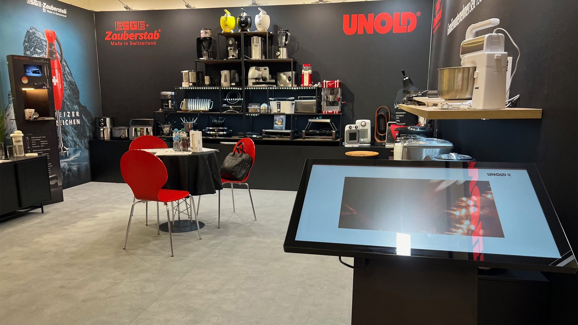 Ein moderner Messestand präsentiert Küchengeräte auf Regalen und Theken, mit einem roten UNOLD-Logo an der Wand, einem Touchscreen-Display davor und zwei roten Stühlen um einen kleinen Tisch.