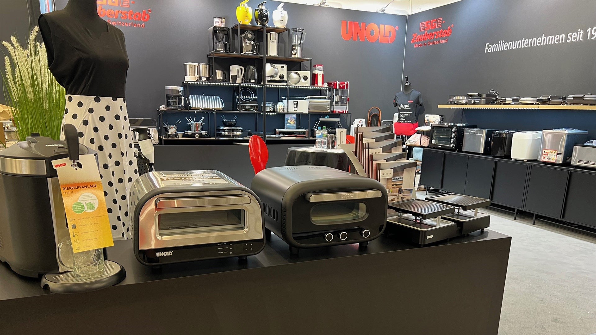 Ein Messestand zeigt auf Tischen und Regalen vor schwarzem Hintergrund mit Markenlogos und Produktbeschreibungen verschiedene Küchengeräte von Unold, darunter Toaster, Grills und Kaffeemaschinen.