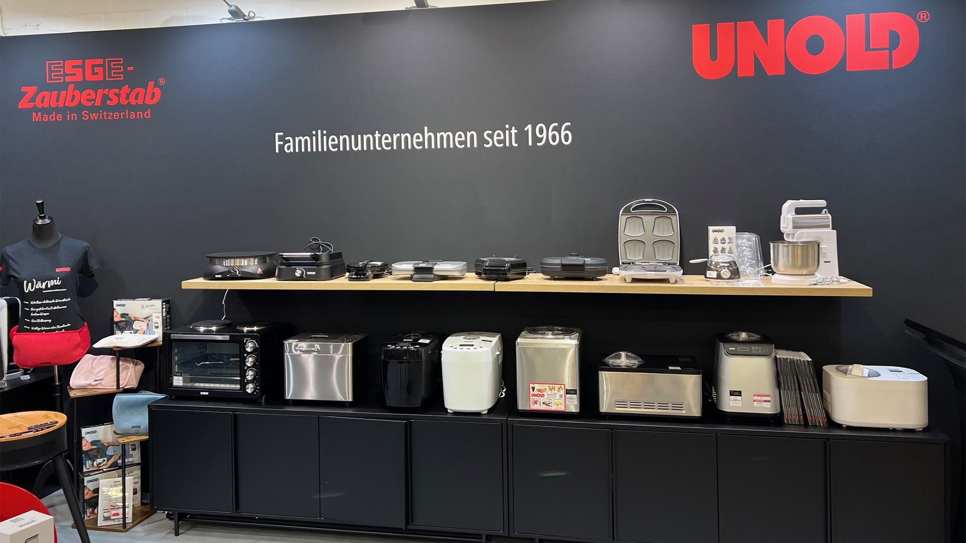 Unter einer schwarzen Wand mit den Logos „ESGE-Zauberstab“ und „UNOLD“ sowie dem Text „Familienunternehmen seit 1966“ sind auf Regalen verschiedene Küchengeräte ausgestellt, darunter Mixer, Toaster, Grills und Brotbackautomaten.