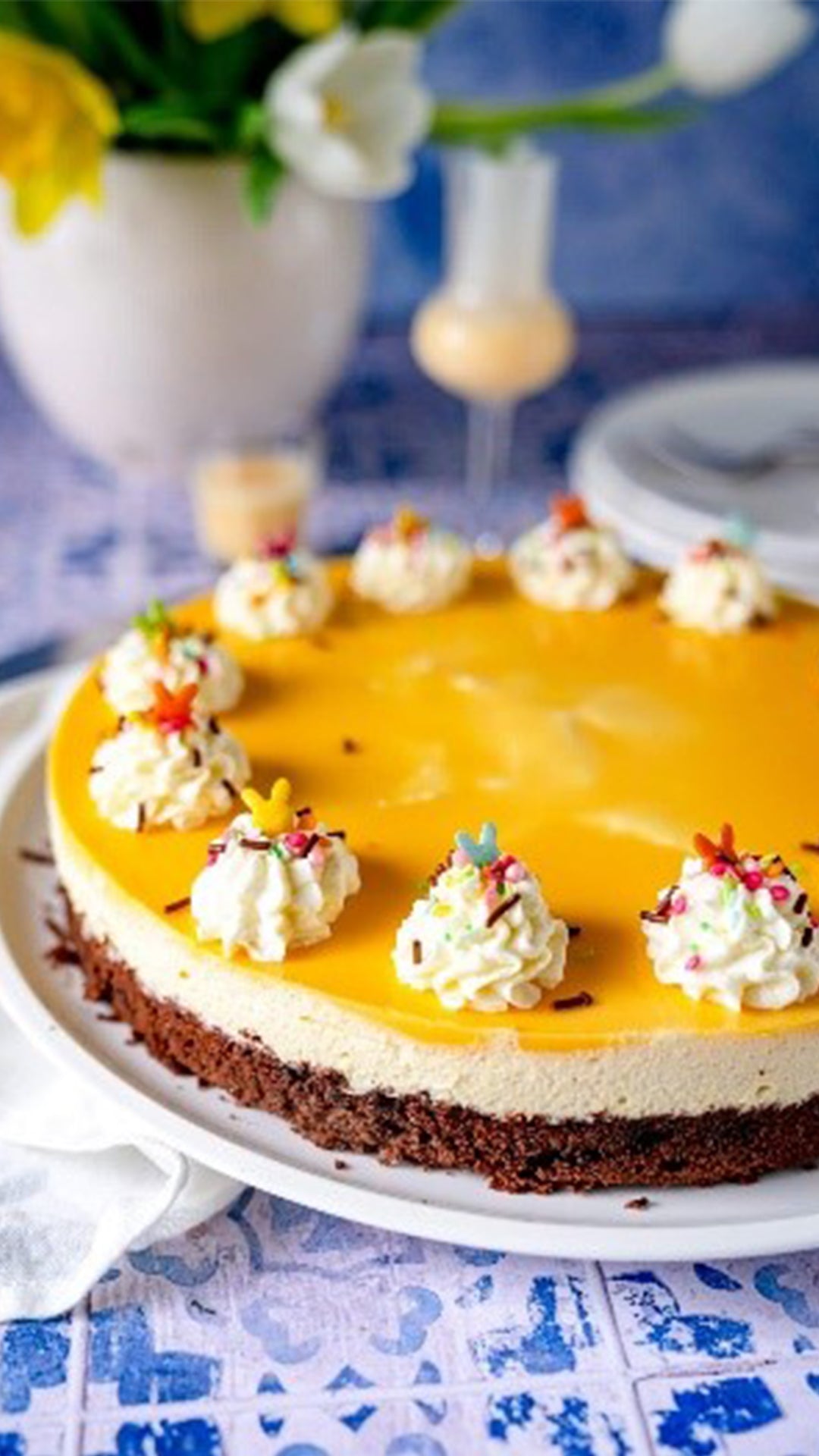 Ein Käsekuchen mit gelber Glasur, Schlagsahne und bunten Streuseln steht auf einem weißen Teller. Im Hintergrund stehen eine Blumenvase, ein Getränk und Teller auf einem blau gemusterten Untergrund.