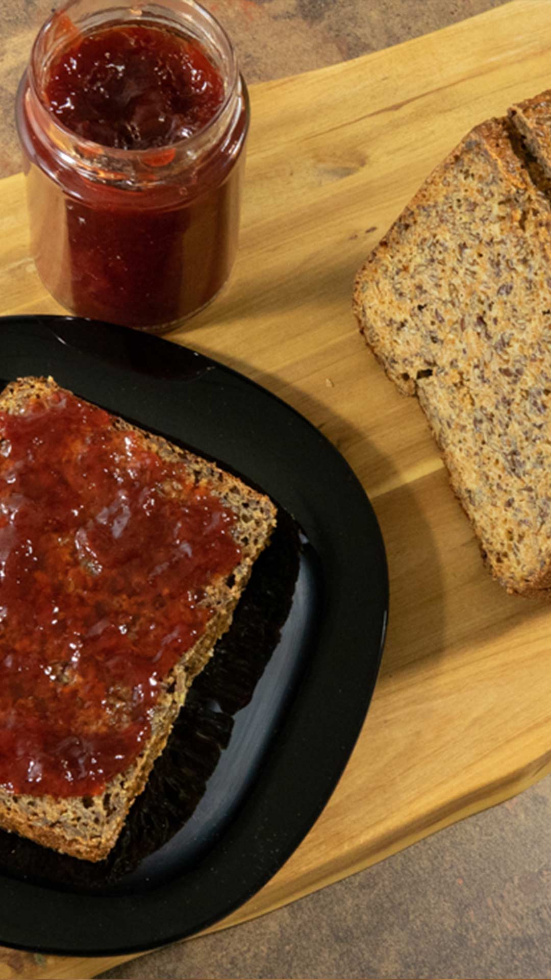 Eine Scheibe Vollkornbrot mit roter Marmelade auf einem schwarzen Teller, daneben ein Glas Marmelade und eine weitere Scheibe Normalbrot, alles auf einer Holzoberfläche.