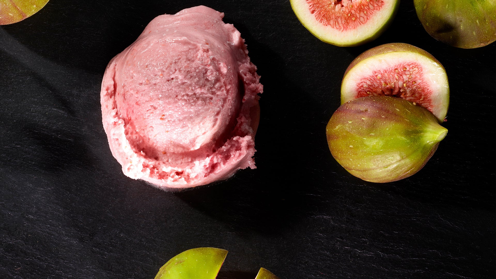 Eine Kugel rosa Eis liegt auf einer dunklen Oberfläche, umgeben von ganzen und halbierten frischen Feigen mit grüner Schale und rosafarbenem Inneren.