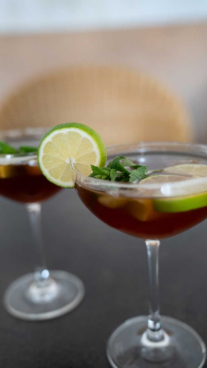 Zwei Cocktailgläser, gefüllt mit einem dunklen Getränk, garniert mit Limettenscheiben und frischen Minzblättern, stehen auf einer dunklen Fläche mit neutralem Hintergrund.