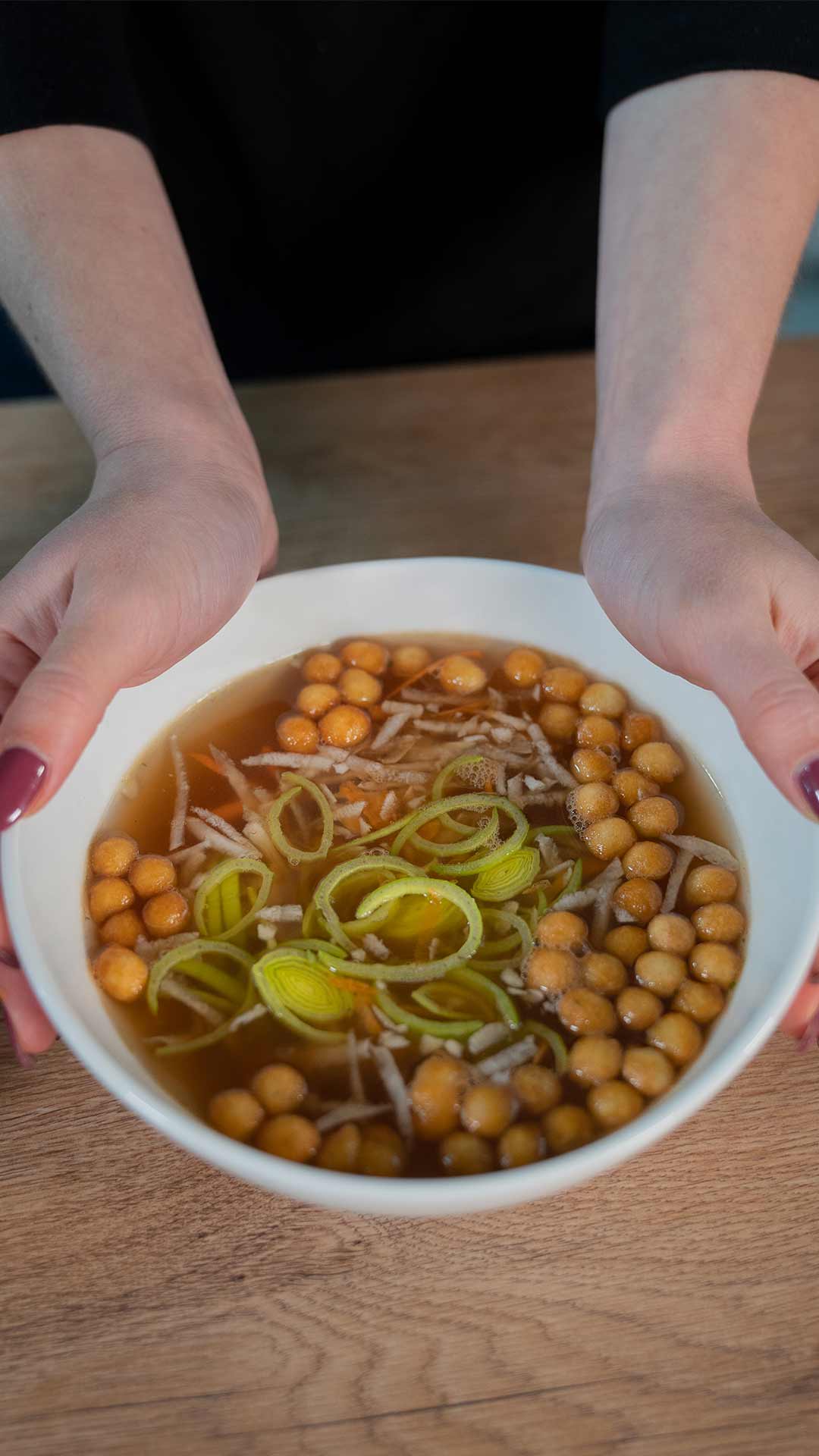 Eine Person hält eine weiße Schüssel mit klarer Suppe, runden Suppenperlen, geschnittenem Lauch und geraspeltem Gemüse auf einem Holztisch.