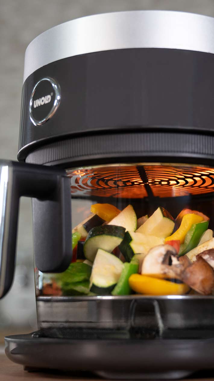 Eine Nahaufnahme einer Heißluftfritteuse, in der verschiedene Gemüsesorten, darunter Pilze, Zucchini, Paprika und Zwiebeln, zubereitet werden, wobei das innere Heizelement orange leuchtet.