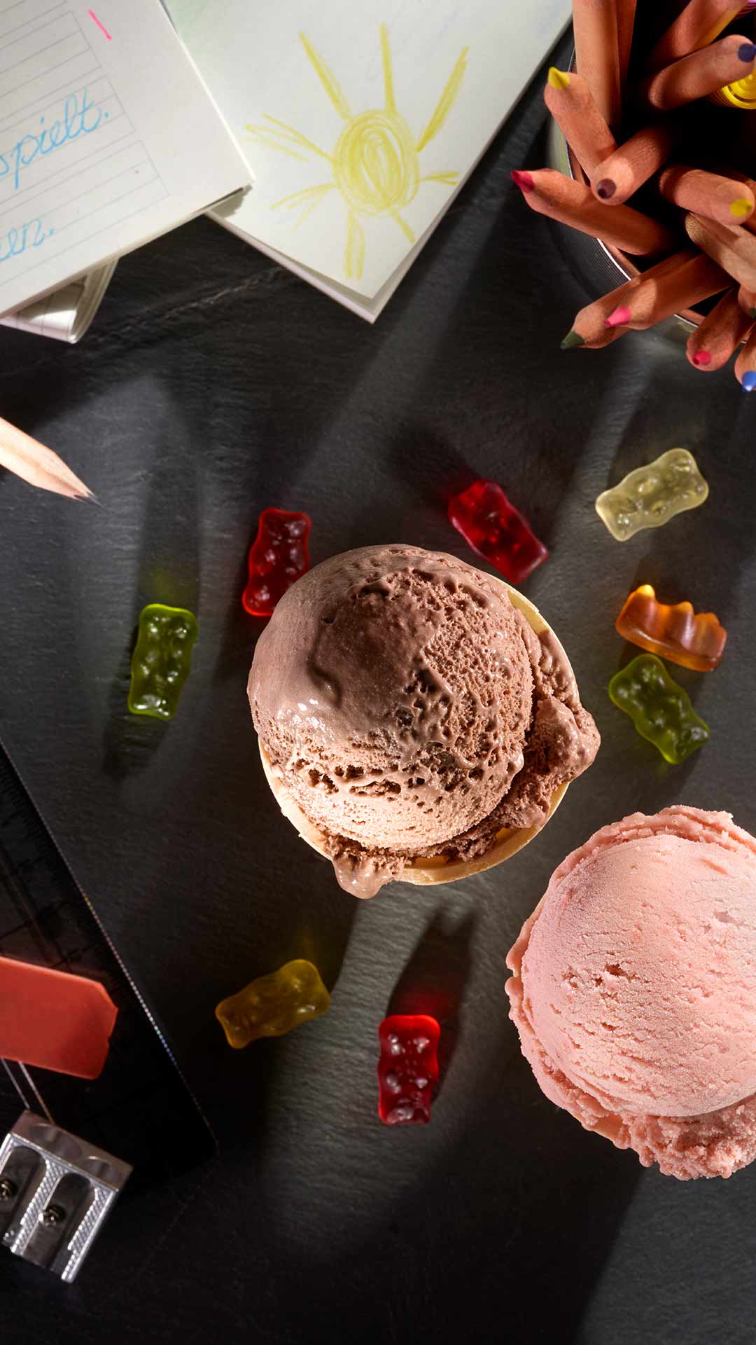 Zwei Kugeln Eis, eine Schokolade und eine Erdbeere, auf einem dunklen Untergrund, umgeben von bunten Gummibärchen, Buntstiften in einem Becher, einer Kinderzeichnung, einem Bleistift und einem Spitzer.