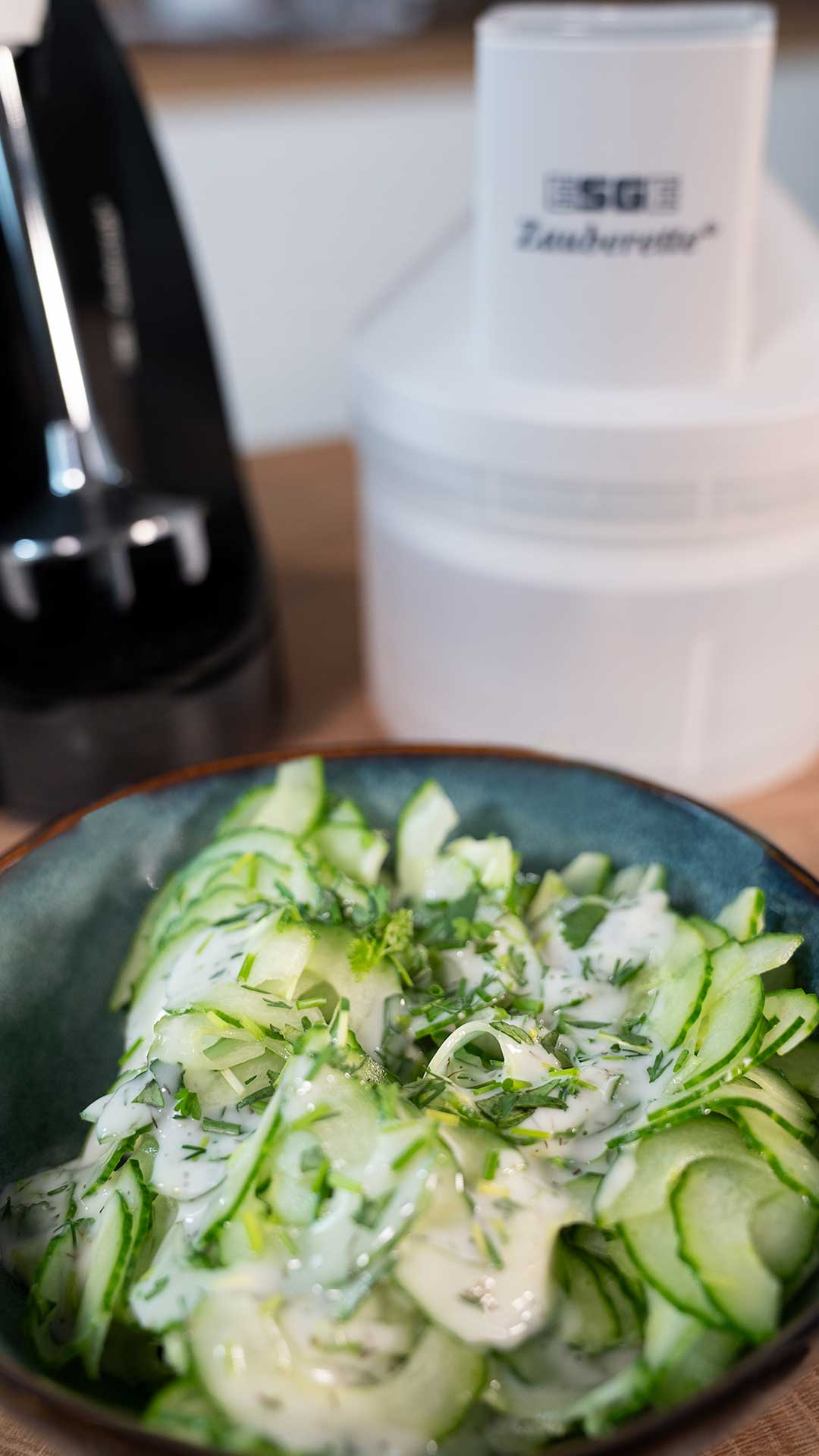 Im Vordergrund steht eine Schüssel Gurkensalat mit cremigem Dressing und frischen Kräutern. Im Hintergrund sind ein Küchengerät und ein weißer Behälter leicht unscharf zu sehen.