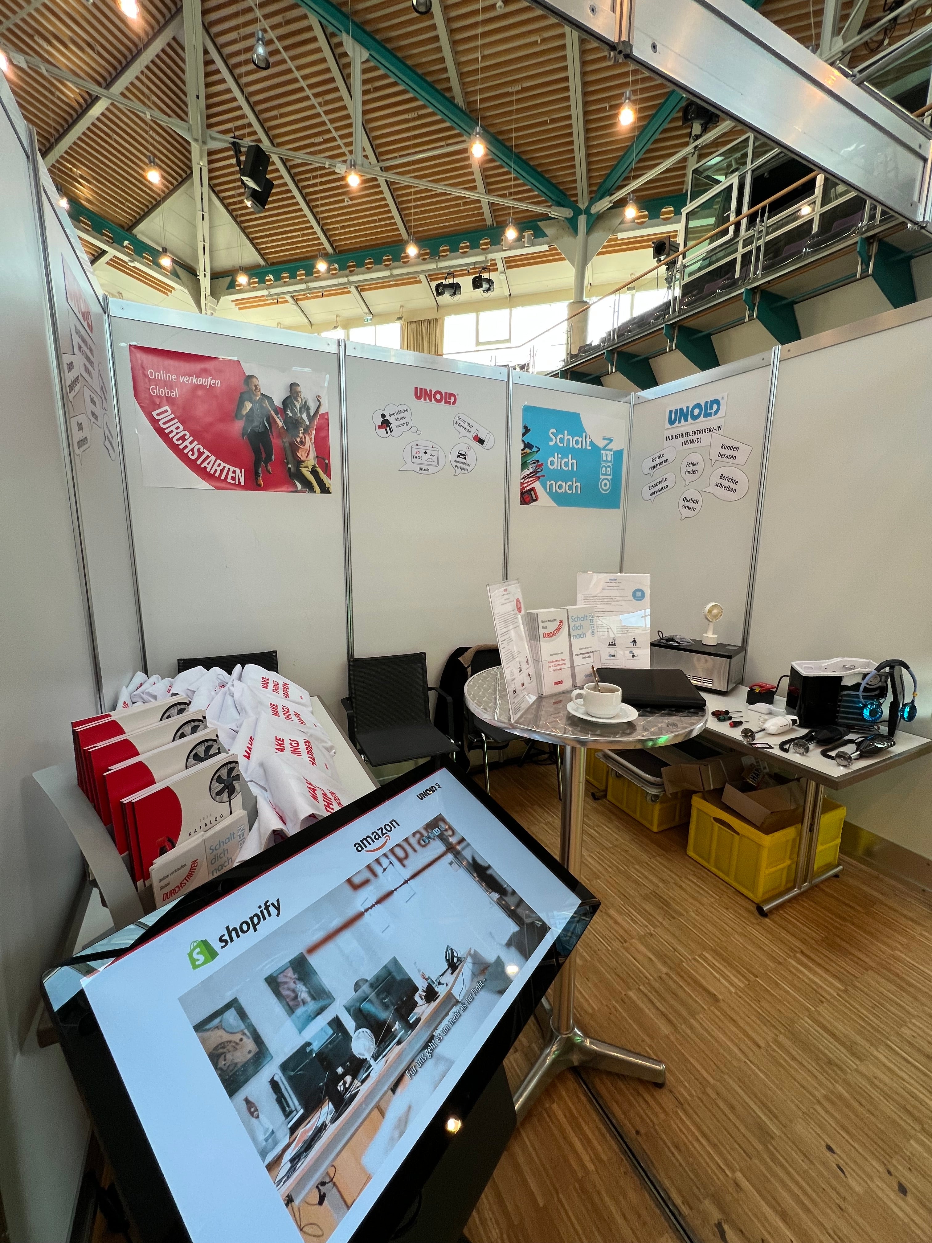 Ein Messestand präsentiert Werbematerialien, einen großen Touchscreen-Monitor mit einer Shopify-Seite, Broschüren, Markentaschen und Produktmuster. Der Stand verfügt über Banner an den Wänden und einen Holzboden.