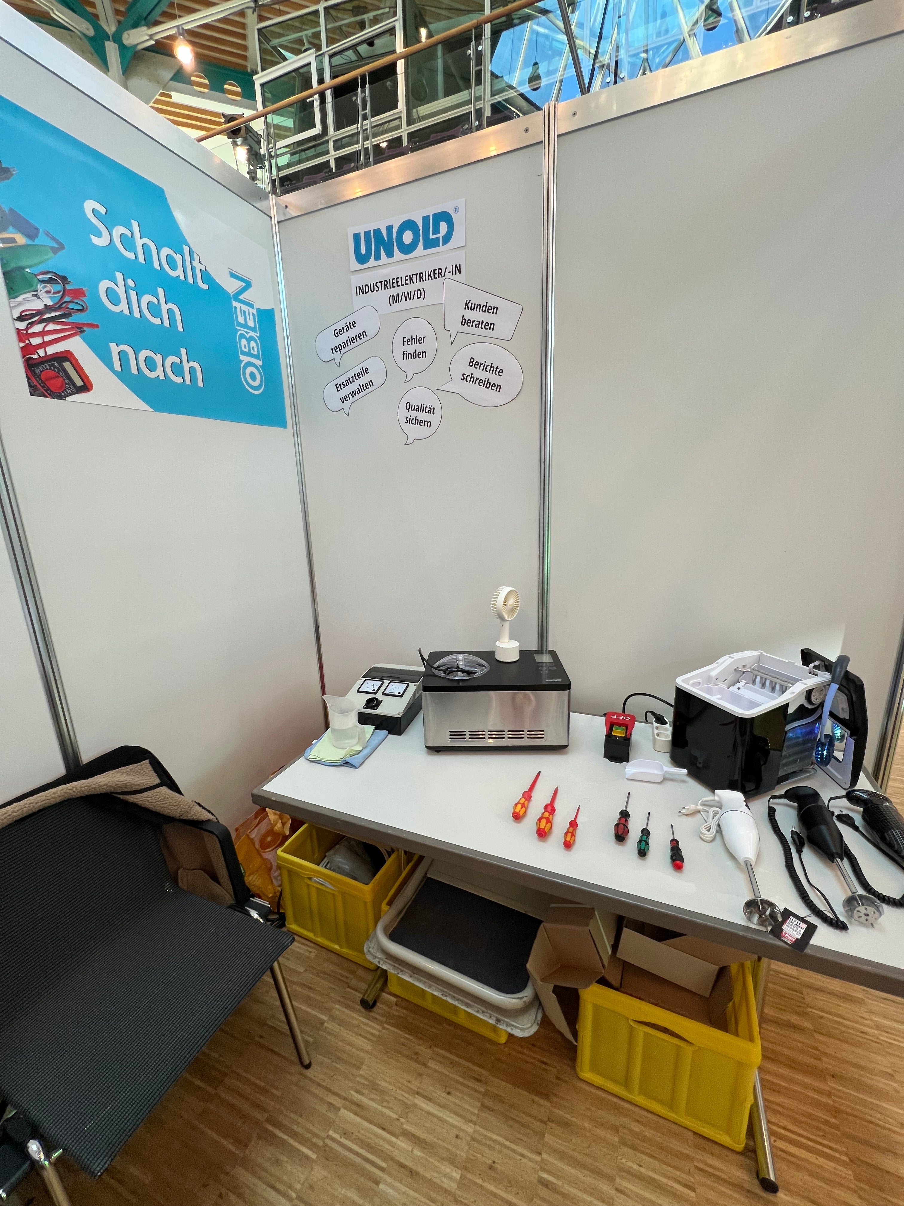 Ein Stand zeigt einen Tisch mit verschiedenen Werkzeugen, Elektrogeräten und kleinen Schraubendrehern. Hinter dem Tisch befinden sich Schilder, darunter eines mit dem UNOLD-Logo. Unter dem Tisch befinden sich gelbe Mülleimer, daneben steht ein Stuhl.