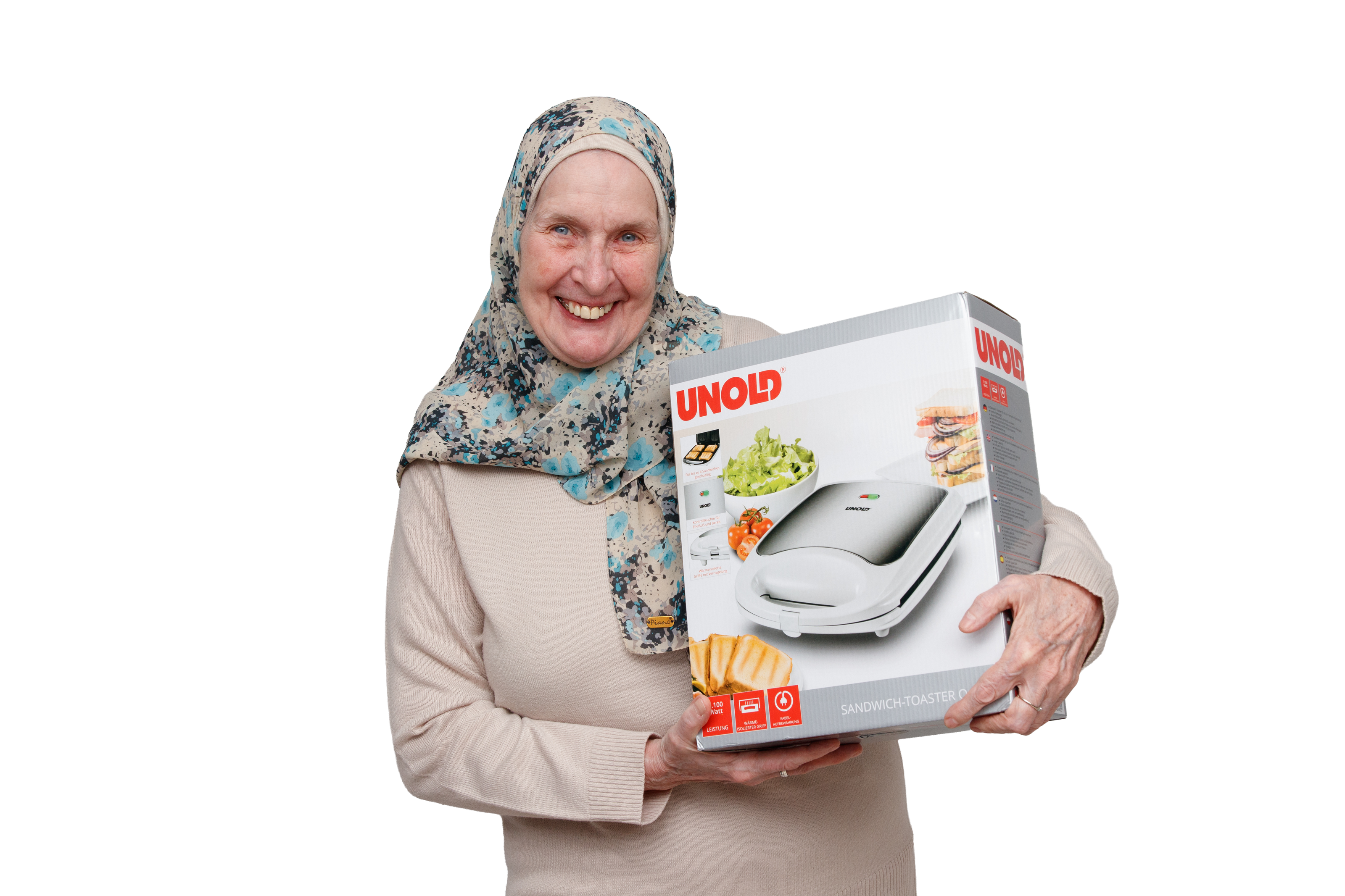 Eine ältere Frau, die einen geblümten Hidschab und einen beigen Pullover trägt, lächelt, während sie einen verpackten UNOLD-Sandwichtoaster vor einem weißen Hintergrund hält.