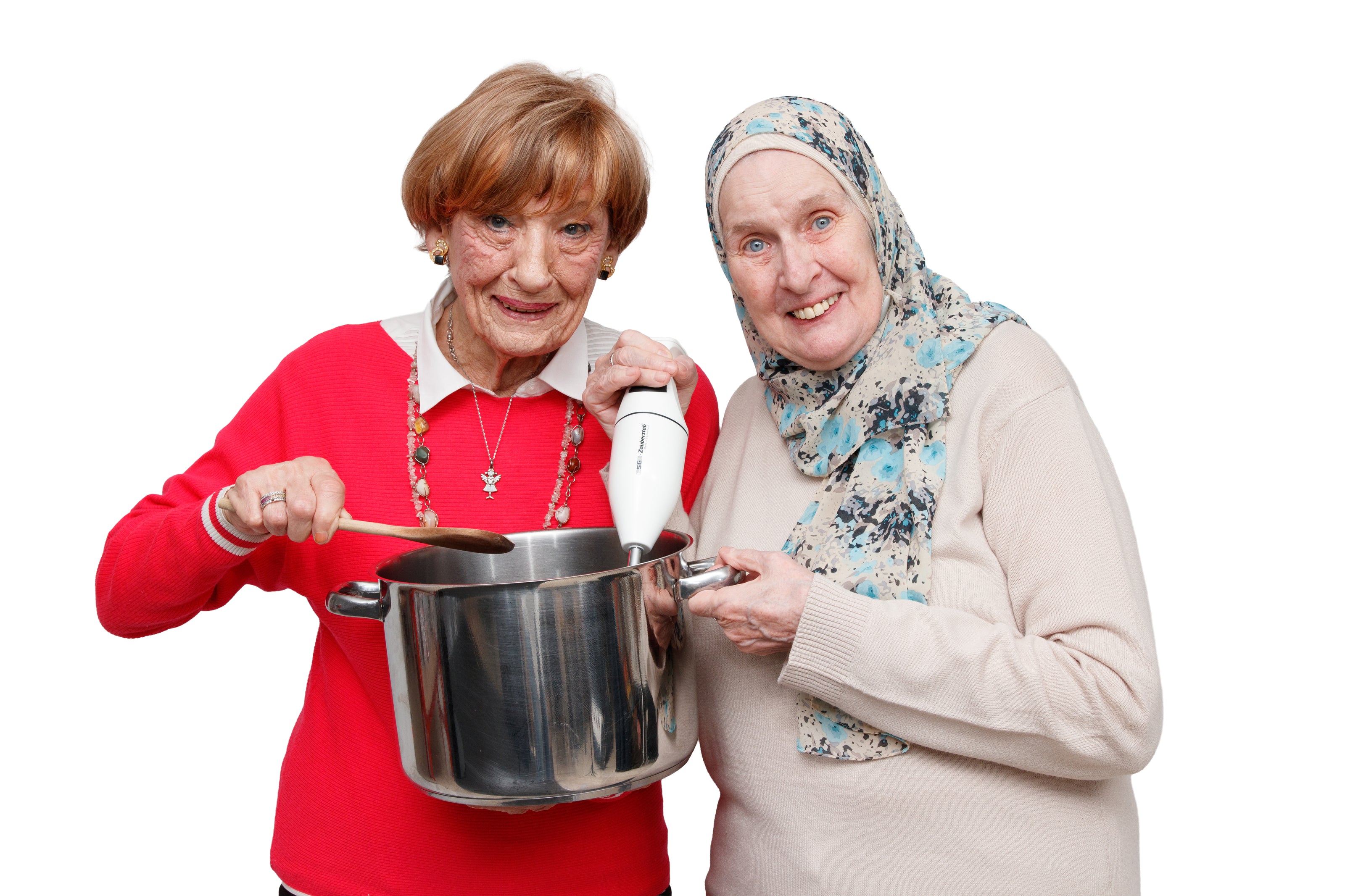 Zwei ältere Frauen lächeln, während sie gemeinsam Essen zubereiten, wobei die eine einen großen Topf mit einem Holzlöffel umrührt und die andere einen Stabmixer benutzt. Beide schauen in die Kamera vor einem weißen Hintergrund.
