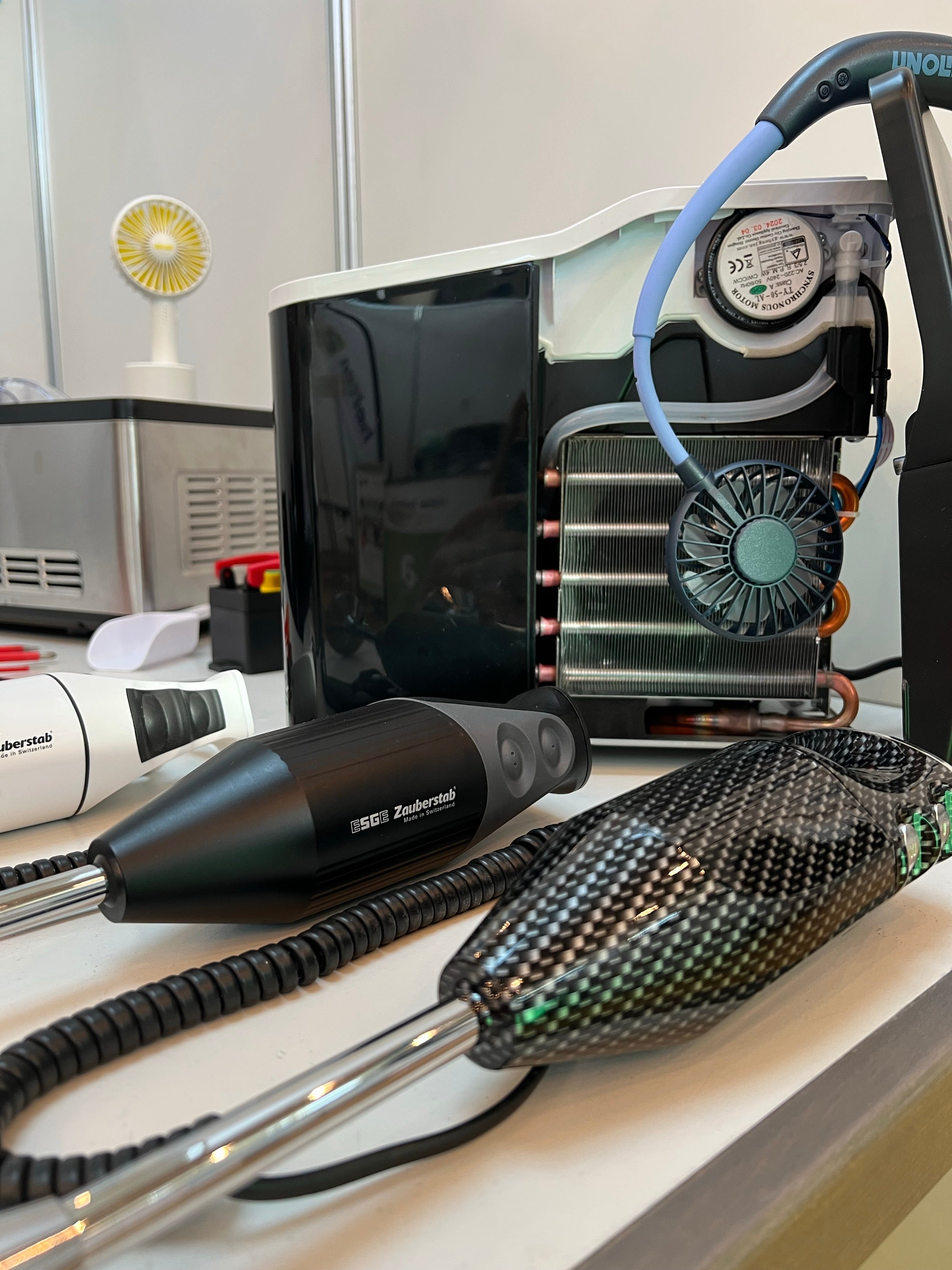 Nahaufnahme moderner Zahnarztgeräte auf einem Tisch, darunter ein Handgerät mit schwarzem Kohlefasermuster, ein schwarzes Gerät mit der Aufschrift „Zephyrtech“ und eine Maschine mit einem Kühlventilator und Spiralkabeln im Hintergrund.