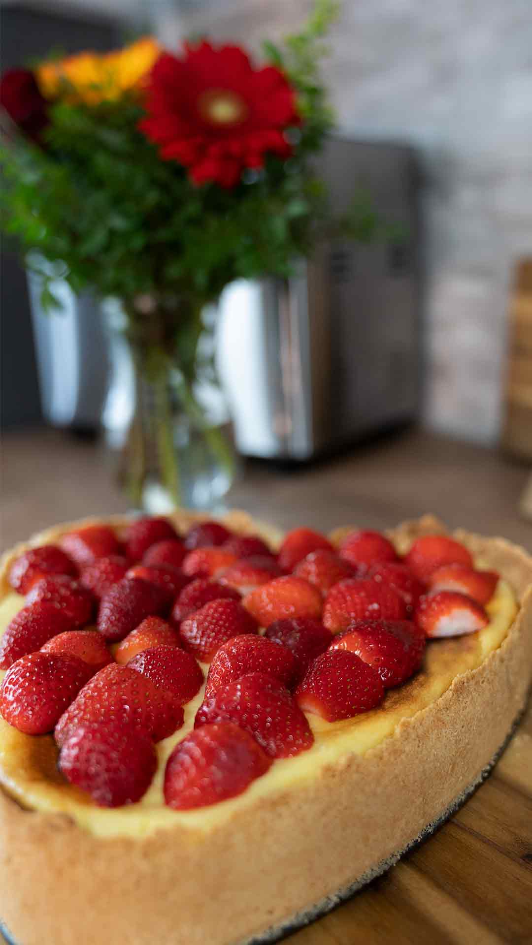 Ein herzförmiger Käsekuchen mit frischen Erdbeeren steht auf einer Holzoberfläche, im Hintergrund sind eine Vase mit Blumen und unscharfe Küchenutensilien zu sehen.