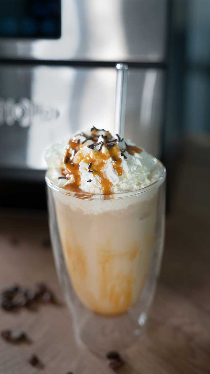 Ein Glas Eiskaffee mit Schlagsahne, Karamell und Schokoladenraspeln sowie einem Strohhalm steht auf einer Holzoberfläche vor einem verschwommenen Küchengerät.