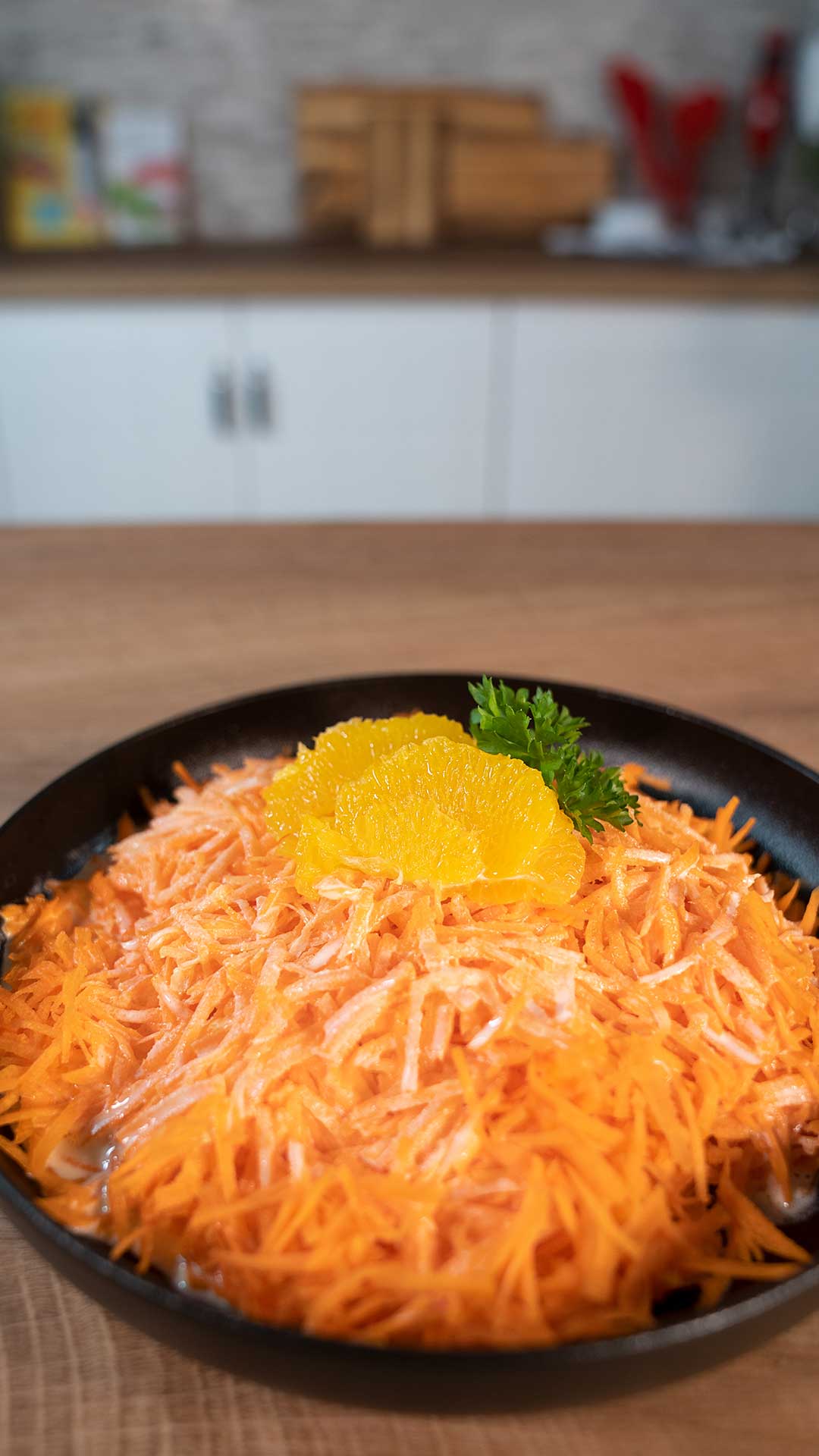 Ein schwarzer Teller gefüllt mit geraspelten Karotten, garniert mit zwei Orangenscheiben und einem Zweig Petersilie, steht auf einer Holzarbeitsplatte in einer modernen Küche.