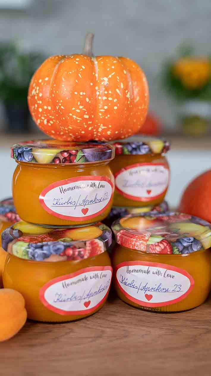 Vier Gläser selbstgemachter Aprikosenmarmelade mit bunten Deckeln und handgeschriebenen Etiketten stehen auf einem Tisch gestapelt. Ein kleiner Deko-Kürbis liegt obenauf. Der Hintergrund ist leicht verschwommen.