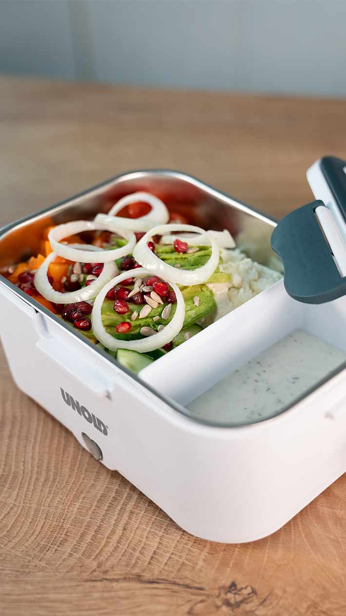 Eine weiße Lunchbox mit einem Metallfach, gefüllt mit Reis, Gurkenscheiben, Zwiebeln, Granatapfelkernen und Sonnenblumenkernen, sowie einem separaten Fach mit einer cremigen weißen Soße. Die Box steht auf einer Holzoberfläche.