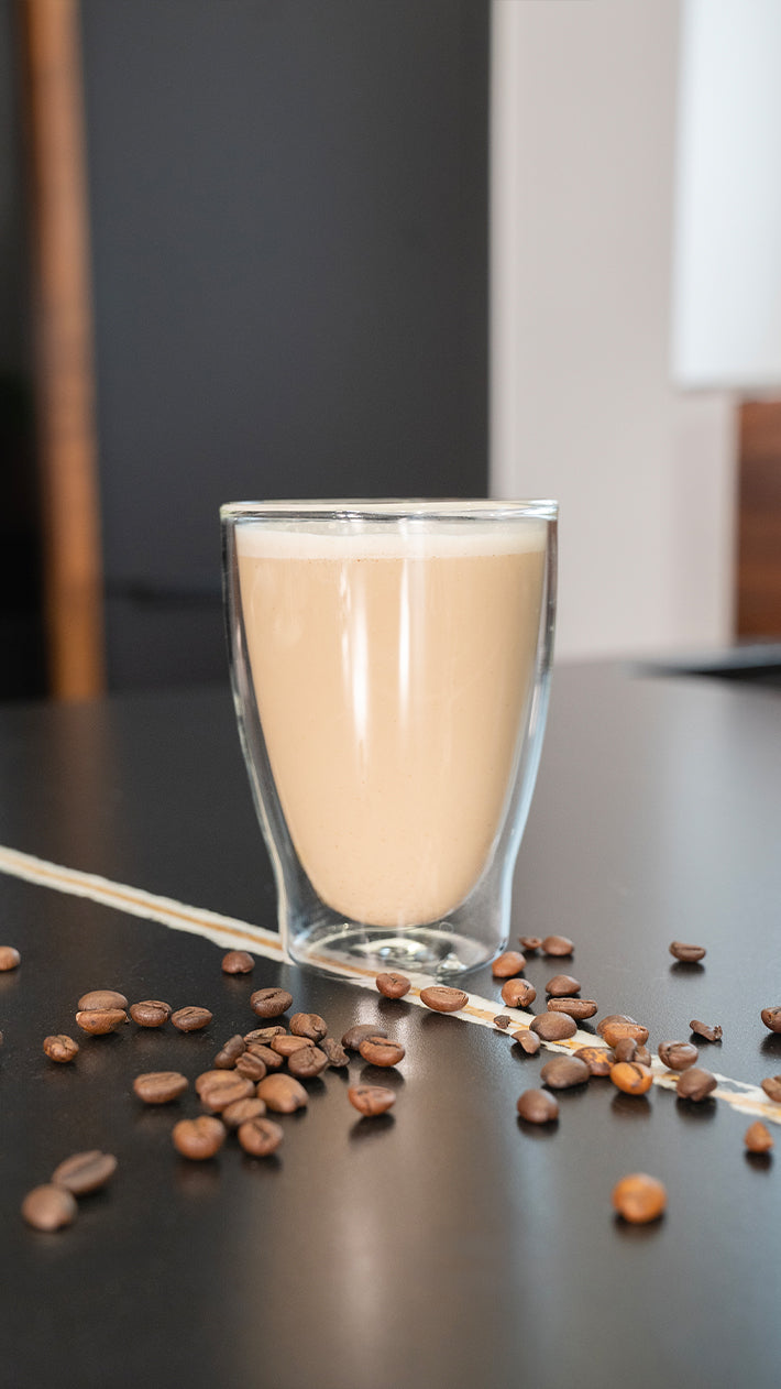 Ein klares doppelwandiges Glas mit schaumigem Kaffee steht auf einem dunklen Tisch, umgeben von verstreuten Kaffeebohnen. Der Hintergrund ist leicht verschwommen.