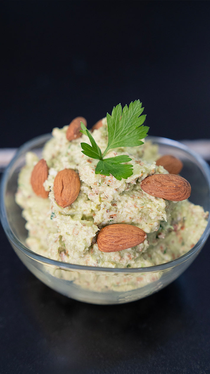 Eine Glasschale gefüllt mit einem cremigen grünen Dip, garniert mit ganzen Mandeln und einem Zweig Petersilie, vor einem dunklen Hintergrund.