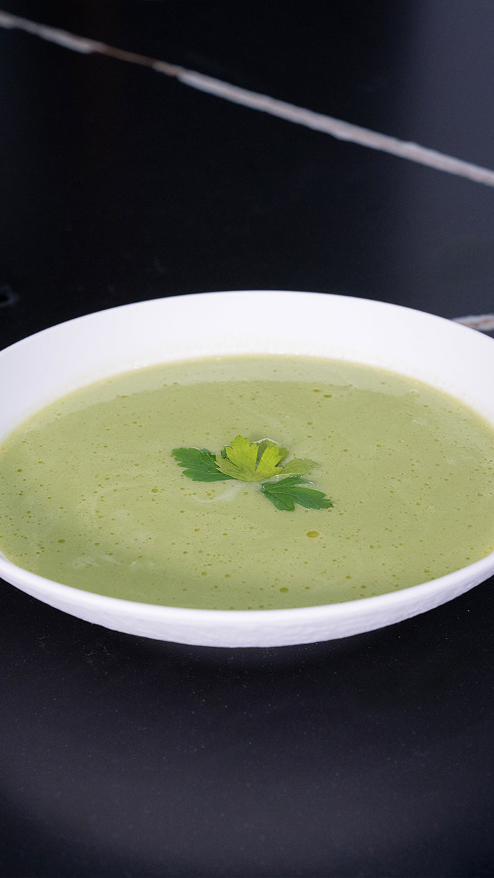 Auf einer schwarzen Fläche steht eine weiße Schüssel mit cremiger grüner Suppe, garniert mit einem Zweig frischer Petersilie.