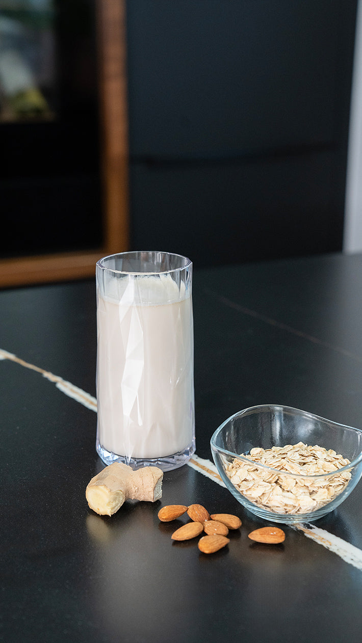 Auf einer dunklen Theke steht ein Glas Pflanzenmilch neben einer kleinen Glasschale mit Haferflocken, einem Stück Ingwerwurzel und mehreren ganzen Mandeln.