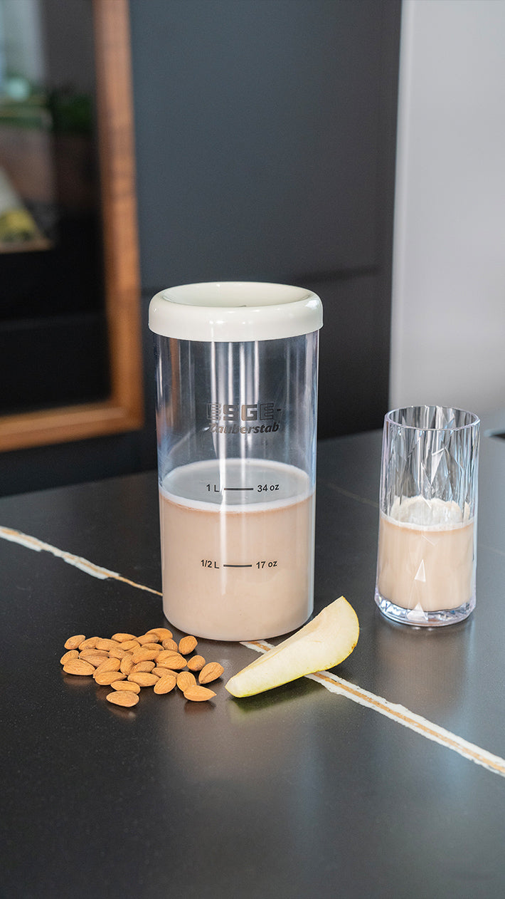 Ein Behälter mit Mandelmilch, ein klares Glas mit Eis, ein kleiner Haufen Mandeln und eine Birnenscheibe stehen auf einer dunklen Arbeitsplatte. Der Behälter ist mit Maßangaben bis 34 oz versehen.