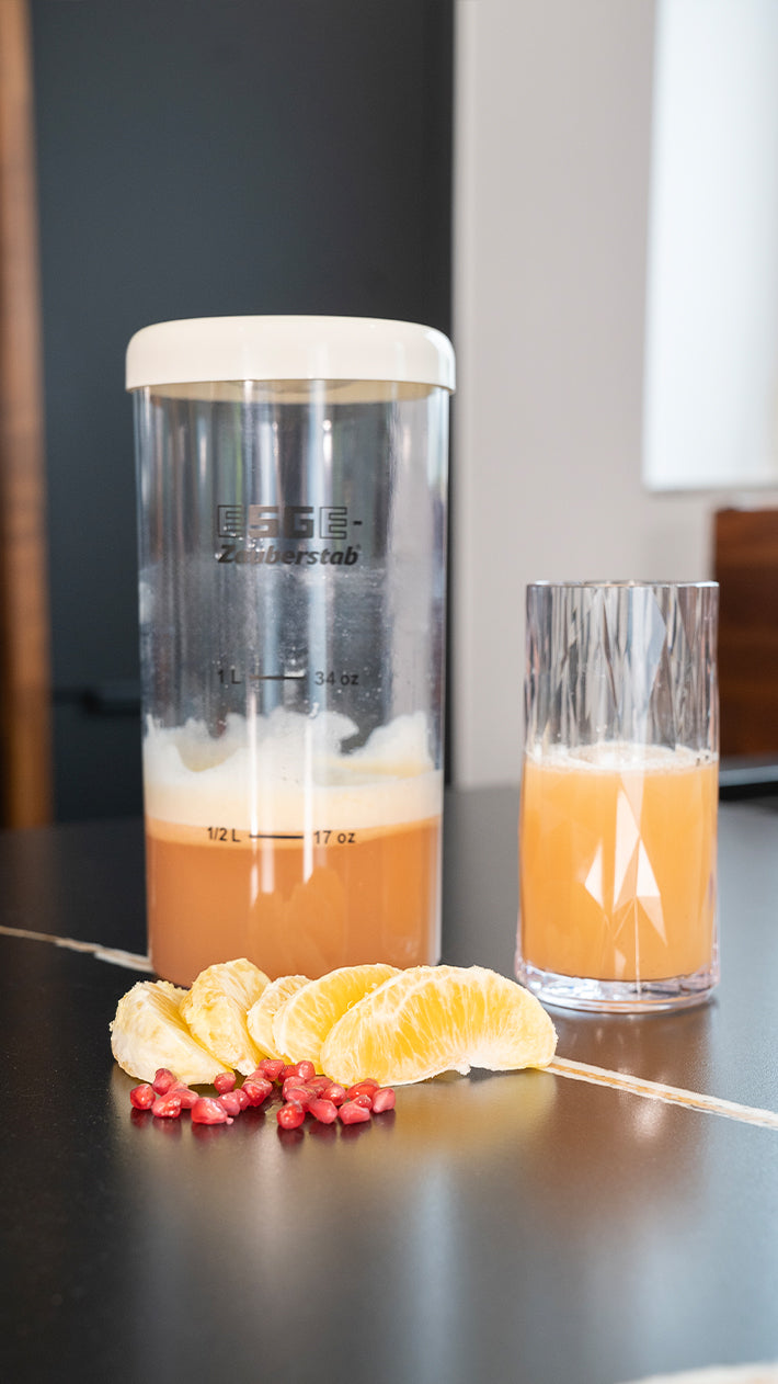 Auf einem Tisch stehen ein Glas und ein Plastikshaker mit hellem Orangensaft, davor liegen verstreut Orangenscheiben und Granatapfelkerne.