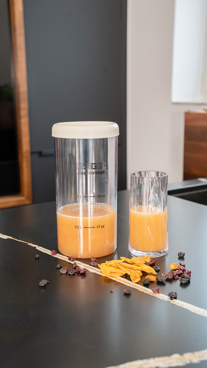 Auf einer schwarzen Marmorplatte stehen ein großer Plastikbehälter und ein Glas, beide mit Orangensaft gefüllt. Daneben liegen getrocknete Früchte und Blütenblätter verstreut.