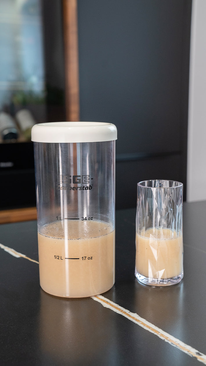 Ein großer transparenter Behälter mit weißem Deckel, der teilweise mit einer hellbraunen Flüssigkeit gefüllt ist, steht neben einem Glas mit einem ähnlichen hellbraunen Getränk auf einer dunklen Arbeitsplatte.