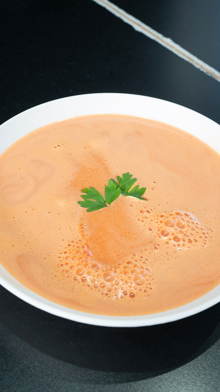 Eine weiße Schüssel gefüllt mit cremiger Orangensuppe, garniert mit zwei kleinen Petersilienblättern in der Mitte, auf einer dunklen Oberfläche.