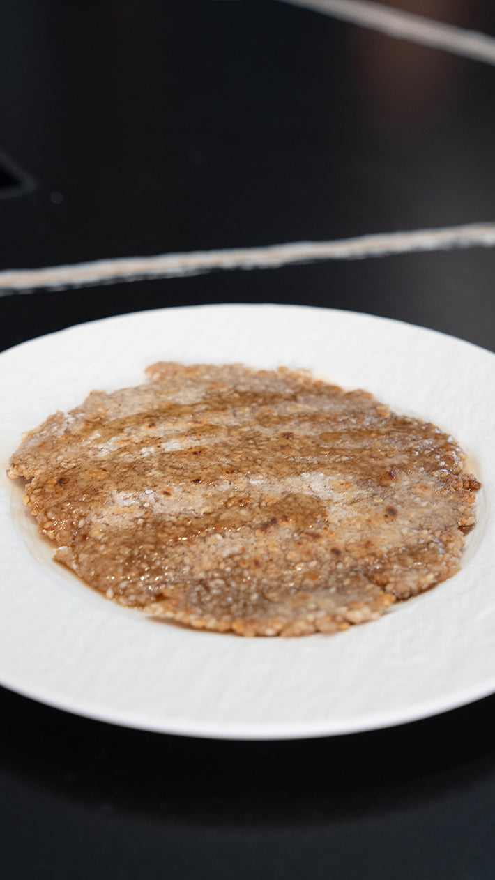 Ein rundes, flaches, bräunliches Lebensmittel mit feuchter und ungleichmäßiger Textur liegt auf einem weißen Teller auf einer dunklen Oberfläche.