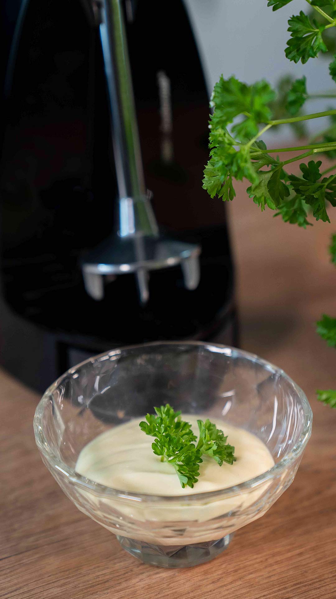 Eine Glasschüssel mit cremiger Sauce, garniert mit frischer Petersilie, steht auf einer Holzfläche. Im Hintergrund sind ein Bund Petersilie und ein Stabmixer zu sehen.
