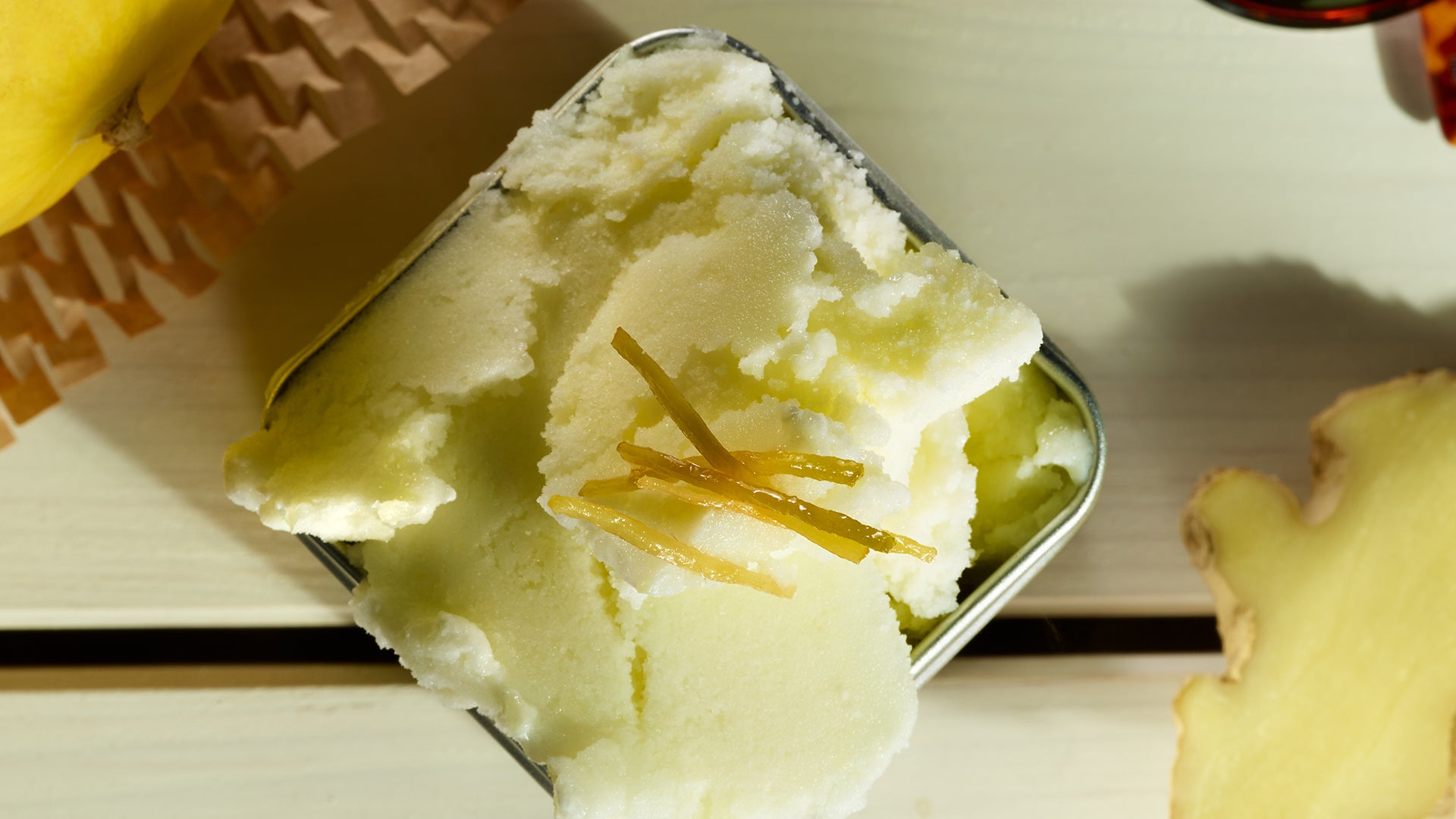 Eine Nahaufnahme eines quadratischen Metallbehälters, gefüllt mit hellgelbem Sorbet, garniert mit dünnen Streifen kandierten Ingwers, auf einer hellen Holzoberfläche.