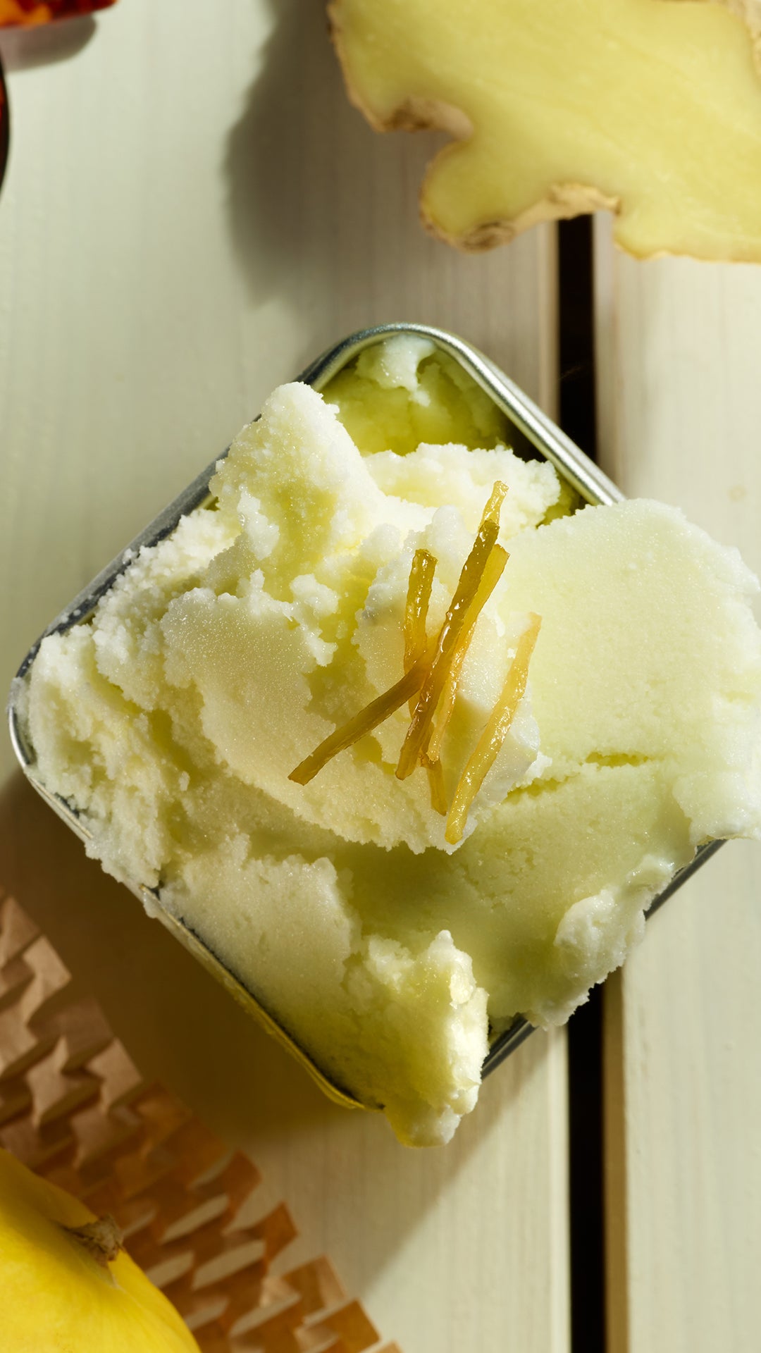 Eine Nahaufnahme von hellgelbem Eis in einer quadratischen Metallschale, garniert mit dünnen Ingwerscheiben, auf einer hellen Holzoberfläche mit einem Stück frischem Ingwer in der Nähe.