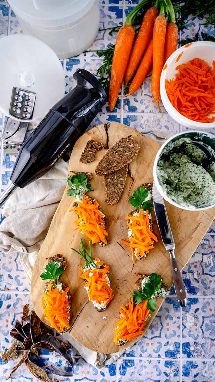 Brotscheiben mit cremigem Aufstrich, geraspelten Karotten und Petersilie auf einem Holzbrett, umgeben von frischen Karotten, geriebenen Karotten, einer Reibe, einem Messer, einem Mixer und einer Schüssel mit Kräuteraufstrich auf einer gemusterten Oberfläche.