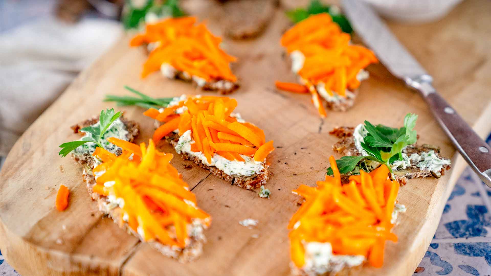 Mehrere Brotscheiben mit Frischkäse, geraspelten Karotten und Petersilienblättern sind auf einem hölzernen Schneidebrett angeordnet, daneben liegt ein Messer.