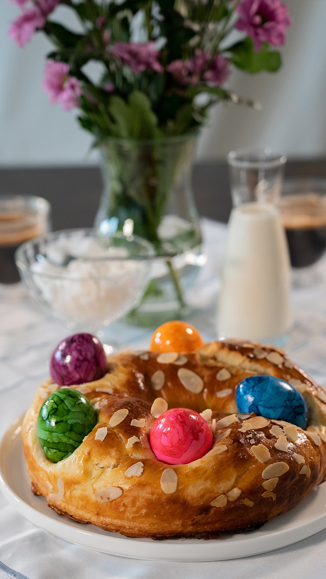 Auf einem Teller liegt ein süßes Brot mit Mandelblättchen und vier bunt gefärbten Eiern, im Hintergrund ein Glas Milch, Schüsseln und eine Vase mit lila Blumen.