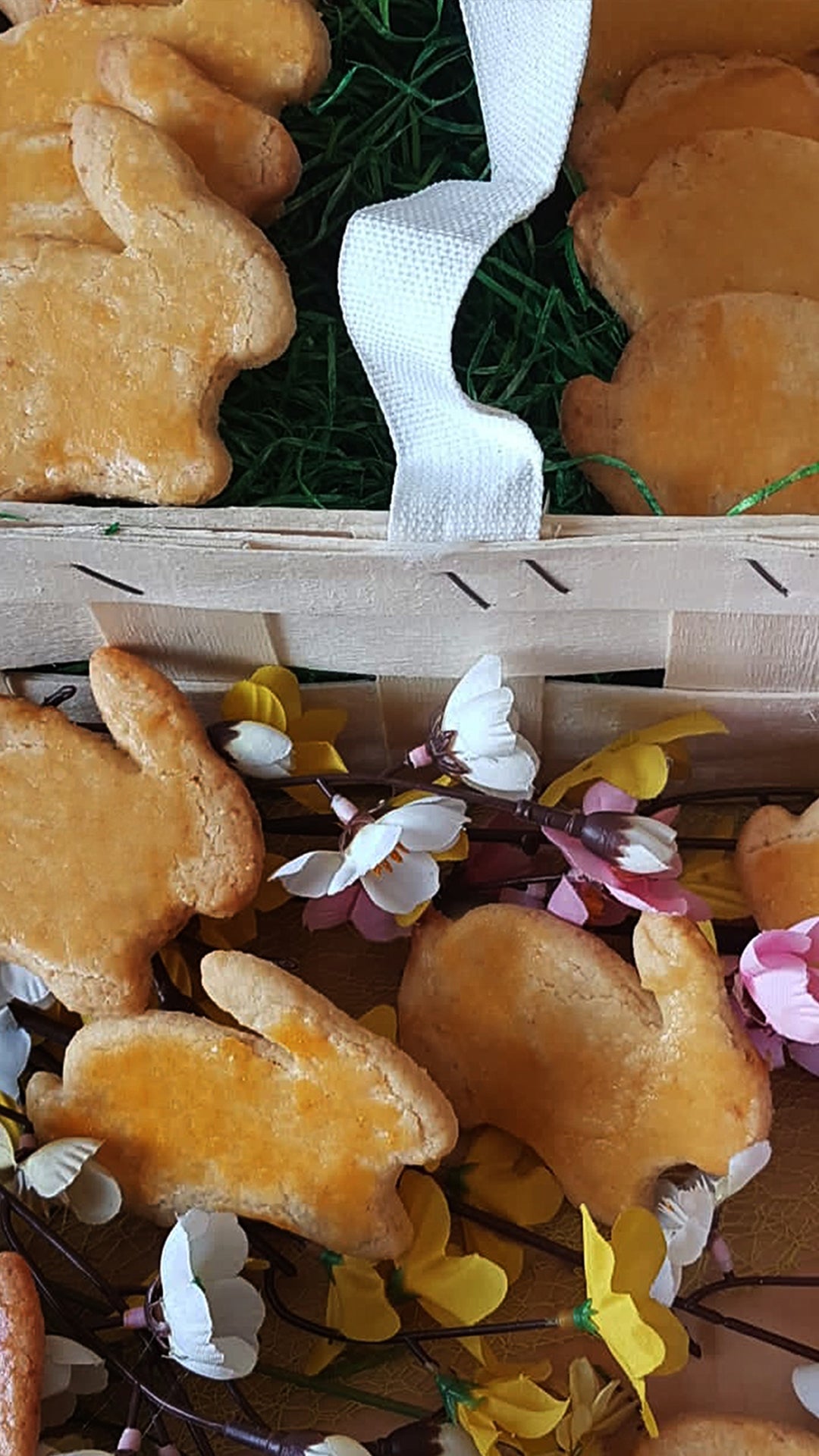 Hasenförmige Kekse sind neben einem dekorativen Korb mit grünem Papierschnipseln und weißem Band angeordnet, umgeben von künstlichen gelben und rosa Blumen.